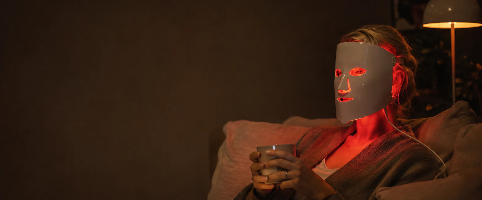 Kvinne slapper av i sofaen en kveld med en LED-maske som lyser rødt og en kopp te i hendene